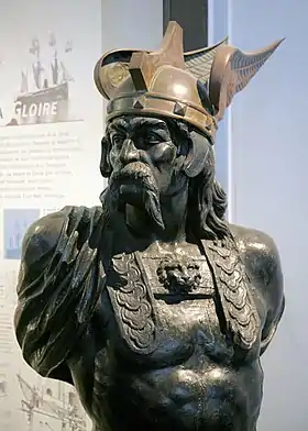 Buste en bronze d'un chef gaulois torse nu, avec une longue moustache et des cheveux longs ainsi qu'un casque celtique de couleur plus claire avec deux ailes latérales.