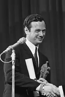 Photographie en noir et blanc d'un homme brun en costume noir souriant devant un microphone et recevant une récompense.