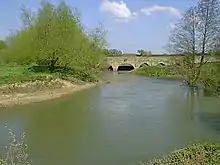 Au premier plan, une rivière aux rives basses avec quelques arbres. En arrière plan un pont de pierre dont on voit 3 arches