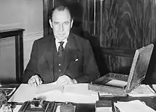 Photographie en noir et blanc d'un homme assis à un bureau et écrivant face à l'objectif.