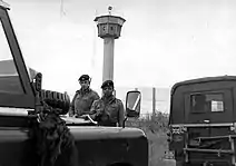Deux soldats britanniques portant des fusils se tiennent derrière deux jeeps dont l'une porte une plaque « British Frontier Service ». Derrière eux se trouve une haute clôture grillagée et une grande tour de surveillance avec un sommet octogonal.