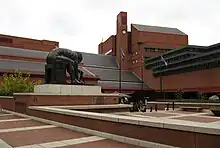 Photographie d'un bâtiment devant une place sur laquelle il y a une statue de bronze qui représente un homme assis qui se baisse devant lui pour toucher le sol.