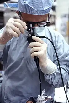 Un homme en tenue stérile tient un fibroscope noir.