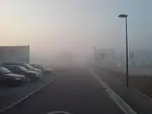 Un épais brouillard matinale envahi une route bordée de parkings.