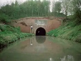 Entrée du tunnel fait de briques rouges au travers de laquelle on peut voir la lumière issue de l'autre extrémité. De chaque coté, il y a des berges herbeuses qui descendent jusque dans l'eau.