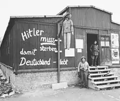 Photo noir et blanc de l’entrée d’une baraque en bois dans le camp de concentration de Buchenwald. Au centre de la photo, près du mur de la baraque, se dresse une potence à laquelle pend un pantin à l’effigie d’Adolf Hitler. Sur le mur de la baraque est peinte l’inscription en allemand : « Hitler muss sterben damit Deutschland lebt ». À droite, un homme, en tenue sombre, se tient debout dans l’encadrement de la porte, au sommet de l’escalier en bois qui mène à l’entrée. Un soldat américain pose debout à côté de l’escalier.