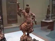 Photo couleur d'une statue sous vitres d'un être de forme humaine en bois marron, le visage emplide fureur et muni d'un sabre et habillé d'une tenue de guerrier.