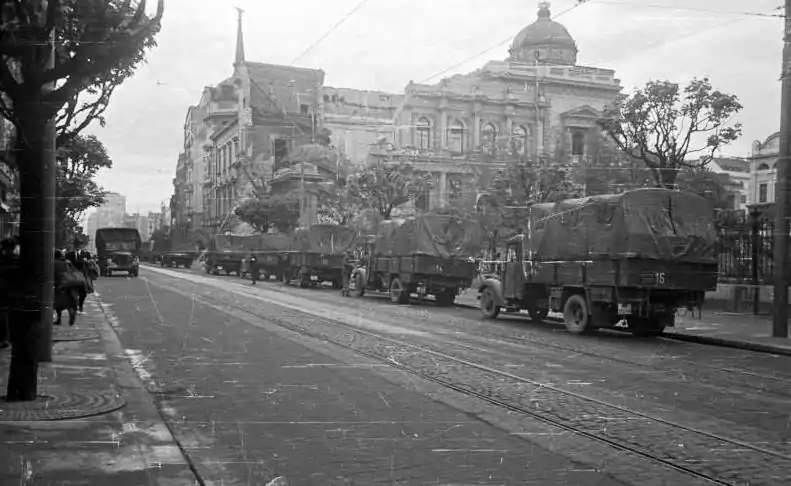 Camions circulant dans une large rue bordée d'arbres avec un grand bâtiment ancien en arrière-plan