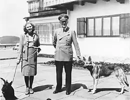 Photographie en noir et blanc d'Adolf Hitler (à droite) et de Eva Braun (à gauche), debout et tenant leurs chiens en laisse, sur la terrasse du Berhof en 1942