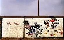 Mur en briques blanches avec, de gauche à droite, le nom de l'album, un trou avec des marteaux qui marchent à l'unisson, des faisceaux lumineux pointant sur une petite personne et un juge gigantesque avec des fesses à la place de la bouche, ainsi que plusieurs autres trous dans le mur avec divers personnages, tels qu'une femme-scorpion.