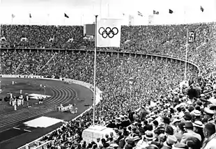 Vue du stade olympique : gradins remplis, drapeaux olympique et nazi visibles.