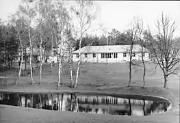 Un bâtiment de plain-pied en fond ; à l'avant-plan, un étang et quelques petits arbres. La photo est en noir et blanc.