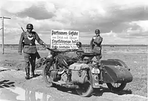 Trois hommes en uniforme, à moto, devant un panneau.