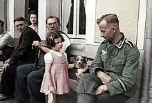 Photo noir et blanc colorisée montrant des civils assis devant une maison avec un soldat allemand tout près d'un chien et d'une fillette