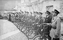 Une douzaine d’hommes, chacun tenant une bicyclette à sa droite, est alignée. Dans le prolongement, en arrière-plan, d’autres policiers juifs se tiennent debout, en rangs.