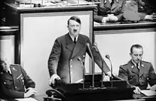 Photo noir et blanc prise le 4 mai 1941, à l’intérieur du Reichstag. Au centre de la photo, Adolf Hitler, debout, mains appuyées de part et d’autre d’un pupitre noir supportant 4 micros, prononce un discours. Il porte une cravate noire, une chemise blanche et une veste croisée sombre. Deux officiers sont assis à ses côtés. L’arrière-plan est constitué de trois panneaux de couleur claire.