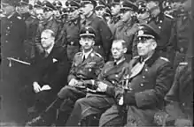 Quatre hommes sont assis devant des rangées de soldats au garde-à-vous. L'homme le plus à gauche est en costume, les autres en uniforme.