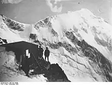 Photo ancienne d'un refuge entouré de trois silhouettes et une montagne enneigée en arrière-plan.