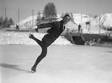 Photographie en noir et blanc d'un patineur au centre de la glace, le corps penché vers l'avant, la jambe gauche relevée et tendue vers l'arrière.