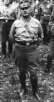 Photographie en noir et blanc représentant Ernst Röhm, en pied, portant l'uniforme de la SA. Derrière lui quelques membres de la SA.