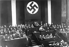 Photographie en noir et blanc prise lors du discours d’Adolf Hitler devant les membres du Reichstag le 23 mars 1933. La moitié supérieure de l’image est dominée par une immense bannière verticale à l’emblème de la croix gammée. Dans la moitié inférieure de l’image, à gauche et à droite, trois rangées de bancs dont les occupants ne sont pas visuellement identifiables. Au centre, de bas en haut, le bureau des sténographes, la tribune, et le siège du président du Reichstag, occupé par Hermann Göring. À la tribune, Adolf Hitler est revêtu de l’uniforme brun des SA ; on distingue à peine le baudrier qui lui barre la poitrine et le brassard à croix gammée à sa manche gauche. Hitler est debout, très légèrement penché vers l’avant ; il a les bras écartés et saisit des deux mains les bords du pupitre.