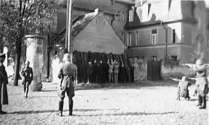 Photographie en noir et blanc montrant, à gauche des hommes adossés en ligne à la façade d'un bâtiment, et à droite le peloton d'exécution.