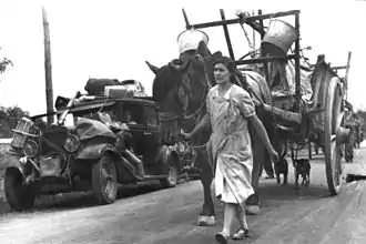 Photo en noir et blanc d'une route avec, allant vers l'objectif, une femme tirant une carriole et, sur le bas-côté, une voiture chargée toute cabossée