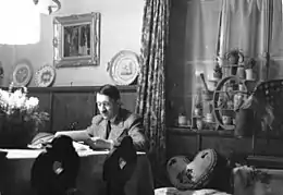 Photo noir et blanc montrant Adolf Hitler, assis à une table, dans une pièce de sa résidence secondaire du Berghof (à Berchstesgaden). L’homme, en costume clair, de face, est en train de lire des documents. L’arrière-plan est divisé par un rideau aux motifs fleuris : à droite, une fenêtre fermée au bord chargé de pots de plantes et, à gauche, un mur blanc supportant un tableau et un meuble en bois d’une couleur foncée sur lequel sont posées, sur la tranche, trois assiettes.