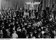 Photographie en noir et blanc d'une foule écoutant un discours d'un orateur placé sous des symboles nazis.
