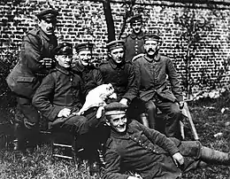 Une photo noir et blanc d'un groupe de sept hommes en tenue militaire de l'armée allemande de 1915. Deux hommes se tiennent debout, devant un mur de briques qui forme l'arrière-plan de la photo, et derrière quatre autres hommes assis dont Adolf Hitler, à droite, reconnaissable à son épaisse moustache. Au premier plan, un septième homme tout sourire se tient allongé sur le côté, dans l'herbe, aux pieds des quatre hommes assis.