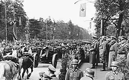 Photo noir et blanc prise le 5 octobre 1939, à Varsovie, en Pologne. Elle montre un défilé militaire de troupes de l’Armée allemande (à gauche) devant Adolf Hitler, debout et bras droit tendu devant lui, avec, dans son dos, une rangée d’officiers (à droite). Des arbres et un ciel clair forment l’arrière-plan de la photo.