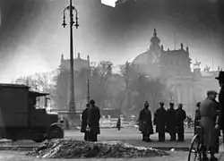 Photo noir et blanc sur laquelle le Reichstag, partiellement caché par une rangée d'arbres, est visible en arrière-plan, le 28 février 1933, lendemain de son incendie. Quelques badauds, en habits sombres, se tiennent debout au premier plan.
