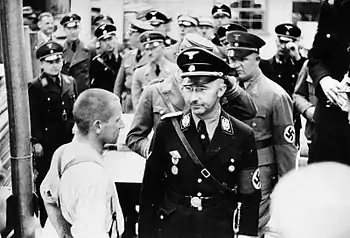 Photographie en noir et blanc représentant Himmler, suivi d'une dizaine de responsables SS, faisant face à un détenu du camp de concentration de Dachau.