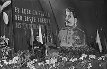 Photo noir et blanc d'un homme en costume sombre prononçant un discours devant un pupitre muni de micros, au premier plan il y a des fleurs et en arrière-plan un portait de Staline en uniforme militaire.