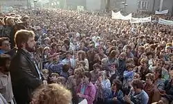 Des milliers de personnes sont rassemblées sur la place d'une ville. Certains portent de larges banderoles et regardent un groupe sur une plateforme située à gauche au premier-plan. Un homme barbu s'adresse à la foule avec des micros.