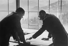 Photo noir et blanc. Adolf Hitler (à droite) et Albert Speer (à gauche), tous deux de profil, en costume sombre, discutent, la tête penchée au-dessus d’une carte étalée sur une plan. En arrière-plan, on voit, à travers une baie vitrée, un paysage de montagne.
