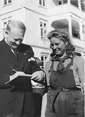 Un homme souriant se penche pour écrire sur un carnet près d'une jeune femme en salopette, visiblement émue.