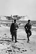 Deux hommes armés, l'un avec un fusil portant une baïonnette et l'autre avec une mitraillette, marchent sur le rivage sous une ligne de crête.