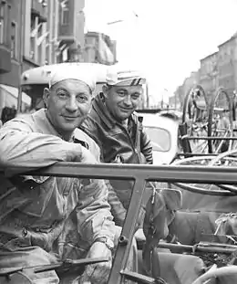 Photo noir et blanc de deux hommes accoudés sur le pare-brise d'une automobile suivant une couse cycliste.