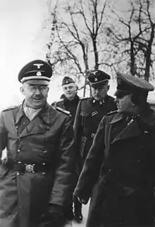 Photographie en noir et blanc représentant Himmler et Eicke, en tenue, marchant côte à côte en extérieur et suivis par deux hommes