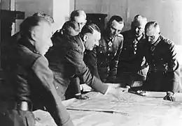 Photo noir et blanc. Dans une pièce aux murs clairs, sept officiers généraux en uniforme suivent les indications données par Adolf Hitler pointant de sa main droite un lieu sur une carte étalée devant eux sur une table. Les huit hommes sont debout autour de la table ; Adolf Hitler et le général Friedrich Paulus sont au centre de la photo.