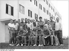 Vingt hommes en uniforme est-allemand prennent la pose pour une photo de groupe devant un bâtiment rectangulaire de couleur claire. La photo est en noir et blanc.