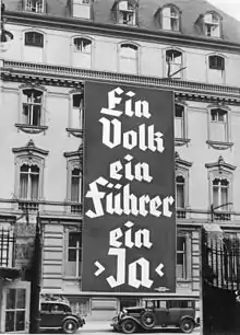 Photo noir et blanc, prise à Berlin en novembre 1933, d'une affiche électorale du NSDAP déployée sur les trois premiers étages de la façade d'un bâtiment de cinq étages (bureau du parti nazi). On peut lire, en grosses lettres, le slogan en allemand : « Ein Volk, ein Führer, ein Ja ».