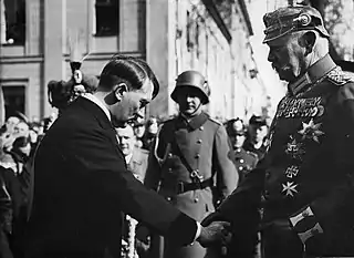 Photographie en noir et blanc prise à Potsdam le 21 mars 1933. À gauche de l'image, Adolf Hitler, vêtu d'une redingote, serre la main du président von Hindenburg, en s'inclinant vers lui. À droite, Hindenburg domine Hitler d'une bonne tête et se tient droit. Ganté, il porte le grand uniforme de maréchal prussien, arborant de nombreuses décorations dont la croix de fer, et un casque à pointe. Au centre et à l'arrière-plan, entre les deux hommes, un militaire avec le lourd casque de combat de l'armée allemande.