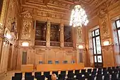 Photographie d'une salle avec en boiserie et dorures et de nombreuses chaises.