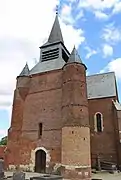 La façade de l'église. À droite une haute tour percée de meurtrières; à gauche une échauguette en brique assise sur un imposant contrefort également en briques.