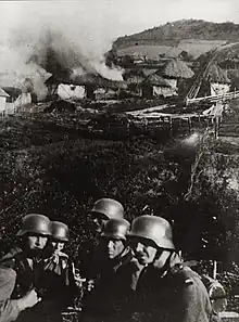 Photo noir et blanc d'un groupe de soldats avec un village en feu en arrière plan.