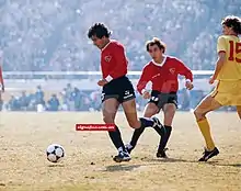 Photographie en couleurs. Un joueur en maillot rouge et short noir court vers la gauche après un ballon, observé par un autre joueur de la même équipe et un autre tout en jaune.