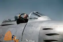 Photographie en couleur d'Aldrin dans le cockpit d'un chasseur à réaction, souriant et saluant le photographe.