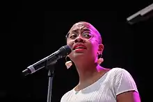 Une femme Noire, crane rasé, des lunettes, des boucles d'oreilles en coquillage, en train de chanter